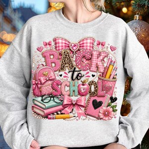 May include: A light grey sweatshirt featuring a graphic print with the text "BACK to SCHOOL". The design includes a pink backpack, stacked books, a pencil holder with pencils, a notebook, and decorative pink bows with leopard print accents.