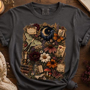 May include: A charcoal gray t-shirt featuring a central graphic print of a crescent moon, a luna moth, a monarch butterfly, various wildflowers, mushrooms, a stack of books, and a candle, all framed within an ornate, vintage-style border.