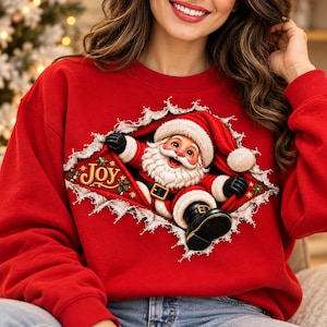 Può includere: Una felpa rossa a girocollo con una grafica di Babbo Natale che sembra emergere da uno strappo nel tessuto. Indossa un abito rosso con cintura nera e bordi bianchi. La parola JOY è stampata in lettere dorate su un nastro rosso.