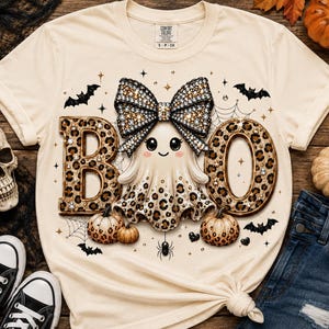 Op de afbeelding: Een cr&egrave;mekleurig t-shirt met een afbeelding van een geest met een grote strik met luipaardprint op het hoofd. De geest staat tussen de letters B en O, die ook een luipaardprint hebben. Het ontwerp bevat kleine pompoenen, vleermuizen en spinnenwebben.