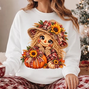 Peut inclure: Un sweat-shirt blanc à manches longues avec une impression graphique représentant un épouvantail aux cheveux blonds, portant un chapeau à carreaux orné de tournesols et de blé. Il tient une grosse citrouille orange avec un tournesol.