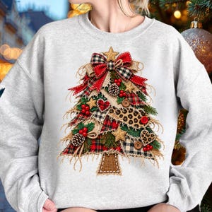 May include: A light gray crewneck sweatshirt featuring a graphic of a Christmas tree constructed from layered fabric strips in red, green, plaid, leopard print, and burlap patterns, accented with red buttons, pinecones, and a bow.