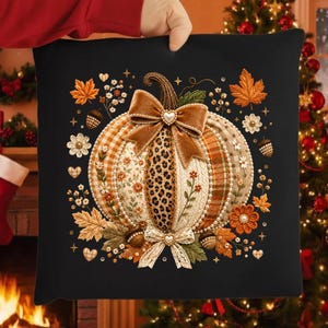 Op de afbeelding: Een zwart vierkant sierkussen met een centrale pompoenafbeelding bestaande uit diverse textuurpanelen, waaronder luipaardprint, bloemmotieven en ruitjes. De pompoen is versierd met een bruine fluwelen strik en een kanten strik met hartvormige accenten.
