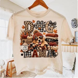 Puede incluir: Una camiseta de color crema con un gr&aacute;fico de un golden retriever que lleva un gorro y gafas de sol en forma de coraz&oacute;n. El dise&ntilde;o incluye calabazas, una taza de caf&eacute; con la inscripci&oacute;n 'Fueled by Coffee and Dog Cuddles' y el texto 'DOG MAMA'.