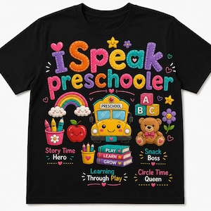 May include: A black short-sleeved t-shirt featuring a colourful graphic with the text "i Speak preschooler". The design includes a yellow school bus, a teddy bear, a rainbow, an apple, crayons, and books labelled "PLAY", "LEARN", and "GROW".