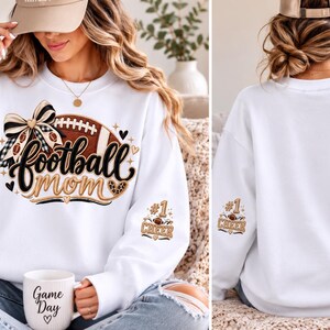 Peut inclure: Un sweat-shirt blanc &agrave; col rond orn&eacute; d'un motif repr&eacute;sentant un ballon de football marron, un n&oelig;ud &agrave; carreaux noir et blanc, et l'inscription "football mom" en lettres noires et dor&eacute;es. La manche porte le texte "#1 CHEER" avec un ballon.