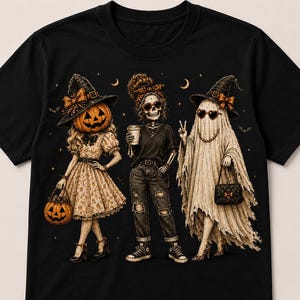 May include: A black t-shirt featuring a graphic of three figures: a pumpkin-headed person in a dress, a skeleton wearing jeans and a t-shirt, and a ghost in a sheet and witch hat. The design includes small stars and crescent moons in the background.