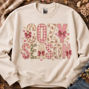 May include: A cream-coloured crewneck sweatshirt featuring the text COZY SEASON in large, decorative letters. The letters are filled with various pink and green floral, gingham, and polka dot patterns, accented with small pink bows and pearls.