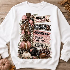 May include: A white long-sleeve sweatshirt featuring a graphic print of a wooden post decorated with a pink and plaid bow, pumpkins, a football helmet, and a pom-pom. Banners display the text Hello Football, Game Day, TOUCHDOWN, Football Mama, and FRIDAY NIGHT LIGHTS.