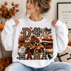 Puede incluir: Una sudadera blanca de cuello redondo con un estampado gr&aacute;fico de un golden retriever con gorro y gafas de sol, rodeado de elementos oto&ntilde;ales como calabazas, hojas y una taza de caf&eacute;. El texto dice DOG MAMA y STAY PAWSITIVE.