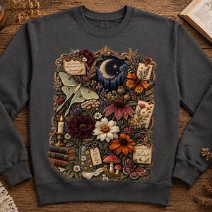 May include: A charcoal gray crewneck sweatshirt featuring a detailed graphic print of a crescent moon, a luna moth, a monarch butterfly, various colorful flowers, a stack of books, a lit candle, and small tags labeled Nocturnal Botanicals.