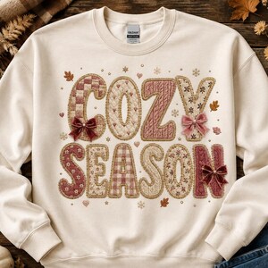 May include: A cream-coloured crewneck sweatshirt featuring the text COZY SEASON in large, textured letters. The letters have various patterns including plaid, quilted, and cable knit designs in shades of pink, brown, and tan, accented with small velvet bows.