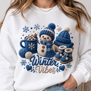 May include: A white sweatshirt featuring a graphic of a snowman wearing a blue knit hat and scarf, a blue mug of hot cocoa with marshmallows, a blue knit hat, and blue knit booties, with the text Winter Vibes in blue and beige script.
