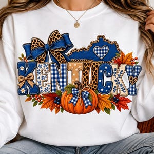 May include: A white long-sleeve sweatshirt featuring a graphic print of the word KENTUCKY in blue and tan patterns. The design includes a blue leopard print bow, a map of Kentucky with a heart, an orange pumpkin, and colorful autumn leaves.