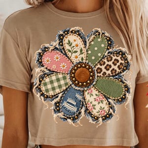May include: A tan short-sleeved t-shirt featuring a large, multi-patterned flower graphic. The flower has a brown center, frayed denim-look petals with gold studs, and various fabric prints including daisies, green polka dots, leopard, plaid, and roses.