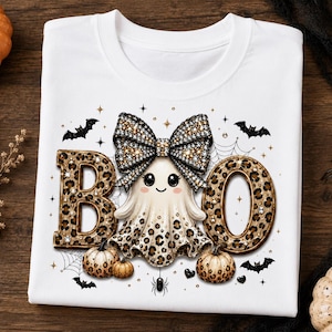 Op de afbeelding: Een wit t-shirt met een grafische print van het woord BOO in een luipaardpatroon. Tussen de letters staat een geest met een grote luipaardstrik. Het ontwerp bevat kleine pompoenen, vleermuizen en spinnenwebben in zwart en goud.