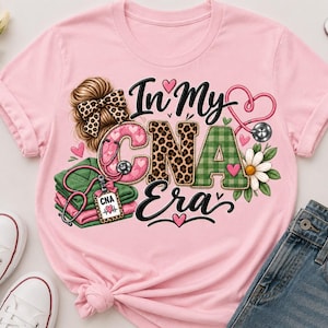 Pode incluir: Uma t-shirt rosa claro com um design gráfico com o texto "In My CNA Era". O design inclui um coque desarrumado com um laço de leopardo, um estetoscópio, uma pilha de uniformes verdes dobrados, um crachá CNA e uma margarida com corações.
