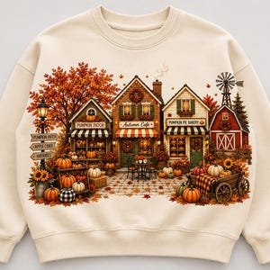 Puede incluir: Una sudadera de cuello redondo color crema con una escena otoñal detallada que muestra un campo de calabazas, un café de otoño y una pastelería de tartas de calabaza. El gráfico incluye árboles naranjas, calabazas, un granero rojo con un molino y letreros que dicen Pumpkin Patch, Apple Cider y Hayrides.