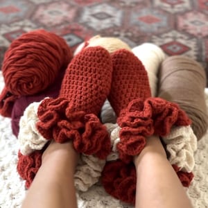 May include: Handmade crochet slippers in a warm red hue, with cream-colored ruffled cuffs. The slippers are surrounded by balls of yarn in red, cream, and brown. A cozy, handcrafted item.