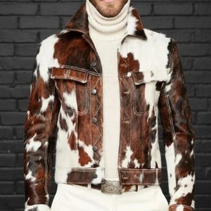 Könnte beinhalten: Eine braun-weiße Kuhfelljacke mit Kragen und Knopfverschluss. Die Jacke hat zwei Brusttaschen und zwei Seitentaschen. Das Model trägt einen cremefarbenen Rollkragenpullover und eine weiße Hose.