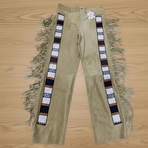 May include: Tan suede trousers with decorative beaded stripes in white, navy, and orange. The trousers feature long fringe along the sides. A tag is attached to the waistband. These trousers are a unique piece of apparel.