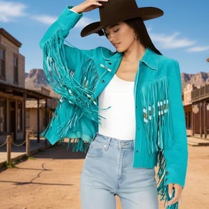 May include: A turquoise suede jacket with long fringe and beaded accents. The jacket has a button-up front and is worn with a white top, light blue jeans, and a brown cowboy hat. The background is a western town.
