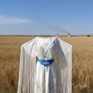 May include: White dress with long fringe sleeves and a blue belt. The dress has a V-neckline with fringe and turquoise accents. The background is a field of wheat under a blue sky.