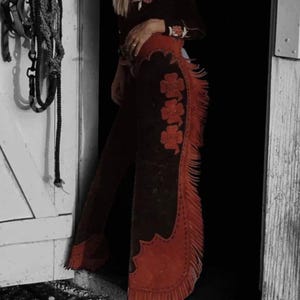 Puede incluir: Chaps de ante marrón con detalles florales rojos y flecos. Los chaps presentan un patrón floral decorativo y flecos rojos en los laterales. Ropa de estilo western.