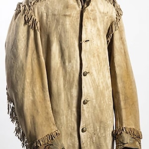 May include: A tan suede jacket with fringe detailing on the shoulders and sleeves. The jacket has a button-up front and decorative stitching on the cuffs. The collar is notched, and the jacket appears to be vintage or antique.
