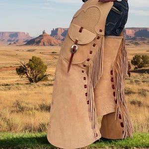 May include: Tan leather chaps with fringe and a pocket, worn over blue jeans. The chaps have decorative stitching and a buckle closure. The background features a desert landscape with mountains and a blue sky.