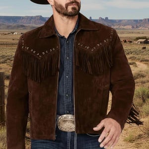 Puede incluir: Chaqueta de ante marrón con flecos y sombrero de vaquero negro. La chaqueta presenta costuras decorativas y ojales metálicos. La persona lleva una camisa vaquera y vaqueros con una hebilla decorativa. El fondo muestra un paisaje desértico.
