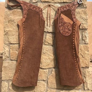 May include: Brown suede leather chaps featuring intricate floral tooling on the upper yokes and a pocket. The garment includes long fringe along the outer legs, a metal buckle closure at the waist, and thin leather ties connecting the two sides.