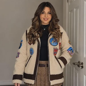 May include: A beige sherpa fleece bomber jacket featuring dark brown trim and multiple colourful embroidered patches on the chest and sleeves. The jacket has a front zip closure and is worn over a black mock neck top and brown plaid skirt.