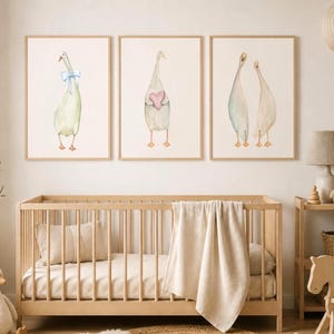 May include: A set of three framed wall art prints featuring watercolour illustrations of geese. The first goose wears a light blue bow, the second holds a pink heart, and the third shows two geese standing together. The frames are light wood.