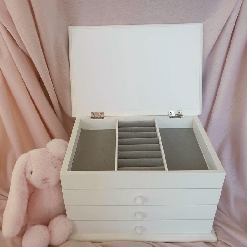 以下が含まれることがあります： 蓋が開いた白い木製のジュエリーボックス。内部はグレーのベルベット張りで、中央のリング収納スペース、2つのサイドコンパートメント、丸い取っ手が付いた3段の引き出しを備えています。隣にはピンクのウサギのぬいぐるみが置かれています。