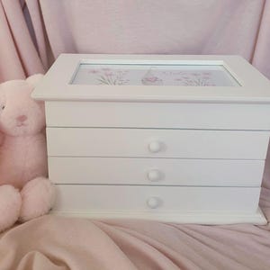 以下が含まれることがあります： 3つの引き出しと丸い取っ手が付いた白い木製のジュエリーボックス。天板のガラスパネルには、ピンクのノームのイラストと「Nela」という名前が描かれています。ボックスの横には、長い耳をした柔らかい淡いピンク色のウサギのぬいぐるみがあります。
