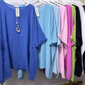 May include: A collection of loose-fitting, short-sleeved tunics hanging on white hangers. The garments are available in various solid colours including royal blue, light blue, pink, magenta, lime green, black, white, brown, and slate blue. Each tunic features a round neckline and side pockets, with a tag reading "MADE IN ITALY" attached to the collar.