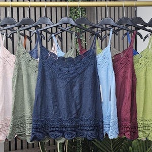 Pode incluir: Uma coleção de blusas sem mangas penduradas num cabide dourado, cada uma com decote quadrado, detalhes em renda bordada e uma bainha festonada a condizer. As peças estão disponíveis em várias cores sólidas, incluindo preto, branco, ferrugem, azul, rosa, verde-azeitona, azul-marinho, azul-claro, bordeaux, verde-lima, cinzento e bege.