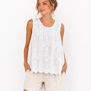 May include: A sleeveless white top featuring vertical panels of floral embroidery and eyelet lace detailing, paired with beige drawstring shorts. The top has a round neckline and a scalloped hemline, while the shorts have a simple, relaxed fit.