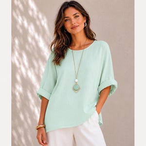 May include: A light mint green short-sleeved blouse with a round neckline and rolled cuffs, paired with white trousers. A long gold-tone necklace features a circular pendant with a matching mint green centre and a white bead accent.