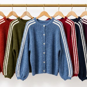 May include: A collection of ten long-sleeved button-down cardigans hanging on wooden hangers. Each cardigan features a solid colour with three white stripes running down the length of the sleeves. The colours include black, grey, red, olive, blue, burgundy, navy, purple, cream, and brown.
