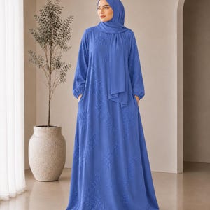 May include: A floor-length cornflower blue dress with long sleeves and a matching hijab. The dress features a subtle tonal floral embroidery pattern throughout the fabric, a round neckline, and side pockets, creating a flowing silhouette.