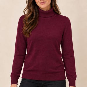 May include: A long-sleeved, deep burgundy roll-neck jumper featuring a ribbed knit collar, cuffs, and hem. The jumper is paired with dark charcoal grey trousers. The garment has a solid colour finish and a fitted silhouette with long sleeves.