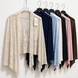 May include: A collection of open-front crochet cardigans hanging on black and white hangers. The cardigans feature a delicate, intricate lace-like pattern and are available in cream, navy blue, light blue, white, black, pink, and dark brown colors.