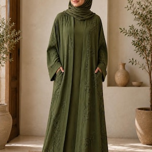 May include: An olive green abaya and matching hijab. The abaya features intricate floral embroidery along the edges and sleeves. The outfit is worn with a matching olive green dress. The person is standing in a room with neutral tones.