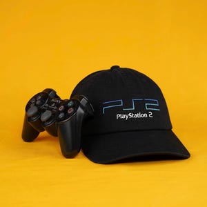 May include: A black baseball cap featuring the embroidered blue and white PS2 logo and the text PlayStation 2 on the front, positioned next to a black wired game controller with analog sticks, buttons, and the Sony brand name printed on it.