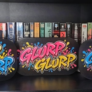 Könnte beinhalten: Drei rechteckige schwarze Schilder mit abgerundeten Ecken, die jeweils das Wort GLURP in einer stilisierten, tropfenden Graffiti-Schrift zeigen. Der Text ist in leuchtendem Gelb, Blau und Pink gehalten und von bunten Farbspritzern umgeben.