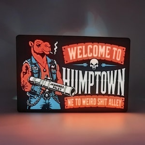 Könnte beinhalten: Ein rechteckiges Leuchtschild mit einem Cartoon-Kamel in einer Jeansweste, das einen Raketenwerfer mit der Aufschrift JASMINES POLE hält. Der Text lautet WELCOME TO HUMPTOWN und NE TO WEIRD SHIT ALLEY mit einem Totenkopf-Symbol.