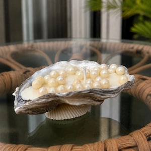 May include: A candle housed within a natural oyster shell, featuring a white wax base embedded with numerous small, round, cream-colored faux pearls and a single centered cotton wick. The shell has a textured, grey exterior and a smooth interior.