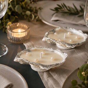 May include: Two oyster shell candles featuring off-white wax poured into natural, irregular shell containers. Each candle has a central white wick and is decorated with several small, round, cream-colored faux pearls embedded in the wax surface.